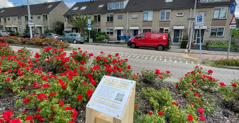 houten bokashi informatie paaltje in plantsoen met rode bloemen. Op de achtergrond een straat met rijtjeshuizen.