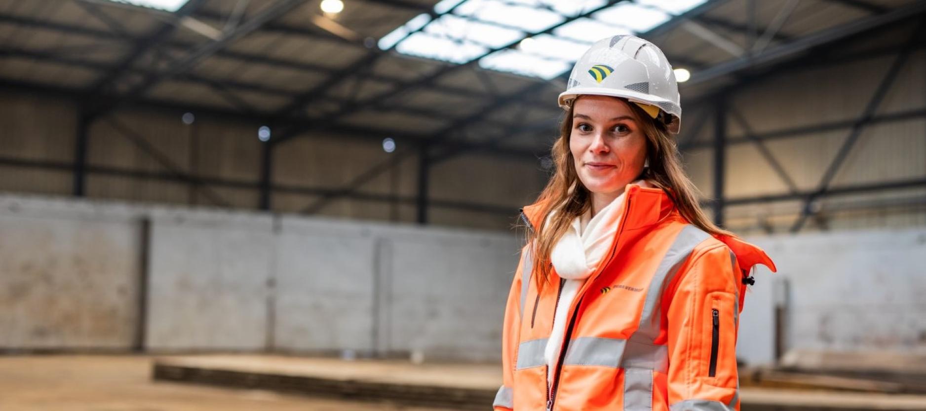 Portretfoto van Sandra van der Lee, gekleed in een oranje bouwjas, witte helm. Sandra staat in een grote lege loods.