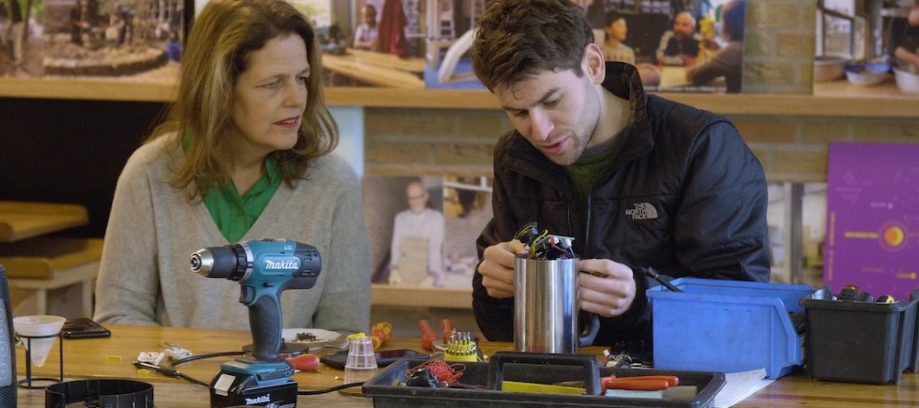 twee mensen zitten achter een houten tafel in het wijkpakijs. Op de tafel staat een boor, hamer en een bak met los gereedschap. de twee mensen kijken aandachtig naar de draden van een elektrische melkopschuimer.