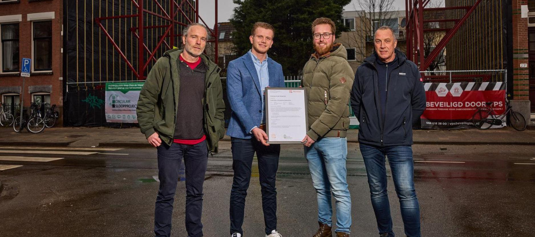 Vier mannen met op de achtergrond de bouwplaats aan de Claes de Vrieselaan. De twee mannen in het midden houden het certificaat vast.