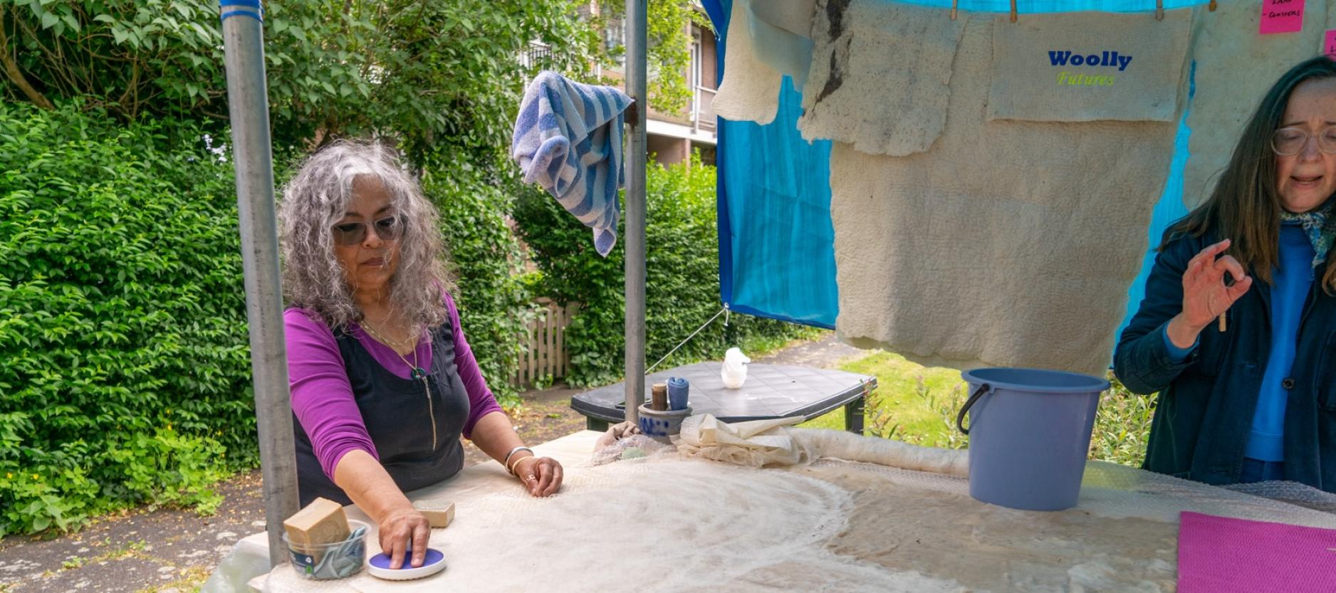 twee vrouwen in een een open tent in de buitenlucht. Groene heg op de achtergrond. Aan een waslijn hangen wollen lappen te drogen. Vrouwen staan een houten tafel. Op de tafel liggen lappen stof en een emmer. Vrouwen zijn bezig met de bewerking van de stof.