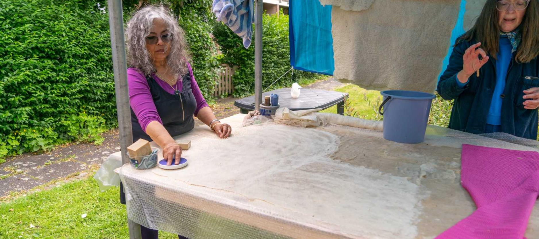 twee vrouwen in een een open tent in de buitenlucht. Groene heg op de achtergrond. Aan een waslijn hangen wollen lappen te drogen. Vrouwen staan een houten tafel. Op de tafel liggen lappen stof en een emmer. Vrouwen zijn bezig met de bewerking van de stof.