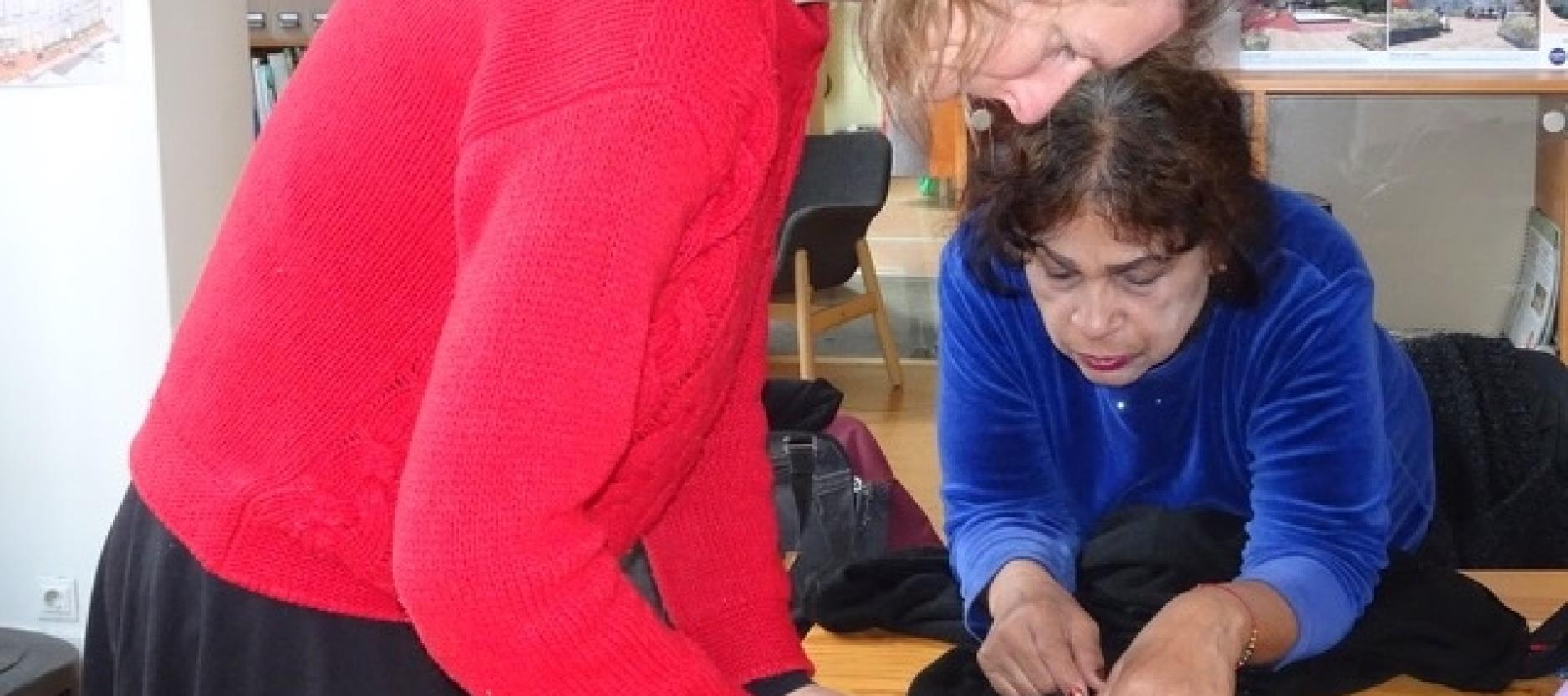 twee vrouwen gebogen over een houten tafel. Ze bekijken een kledingstuk. Op de tafel ligt een schaar en een meetlint.