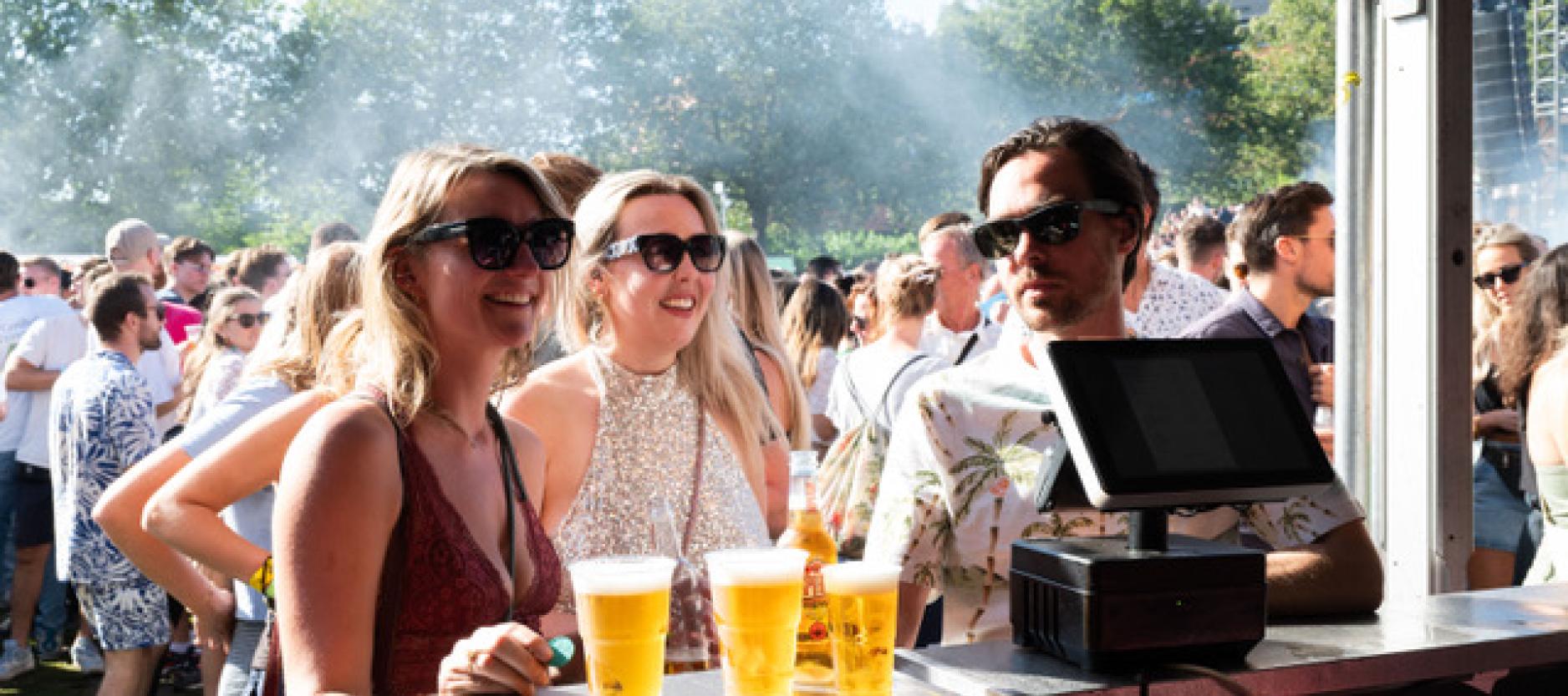 3 festivalgangers staan aan de bar met drie bier op de bar, naast de kassa. Op de achtergrond staan bomen en veel andere festivalgangers.