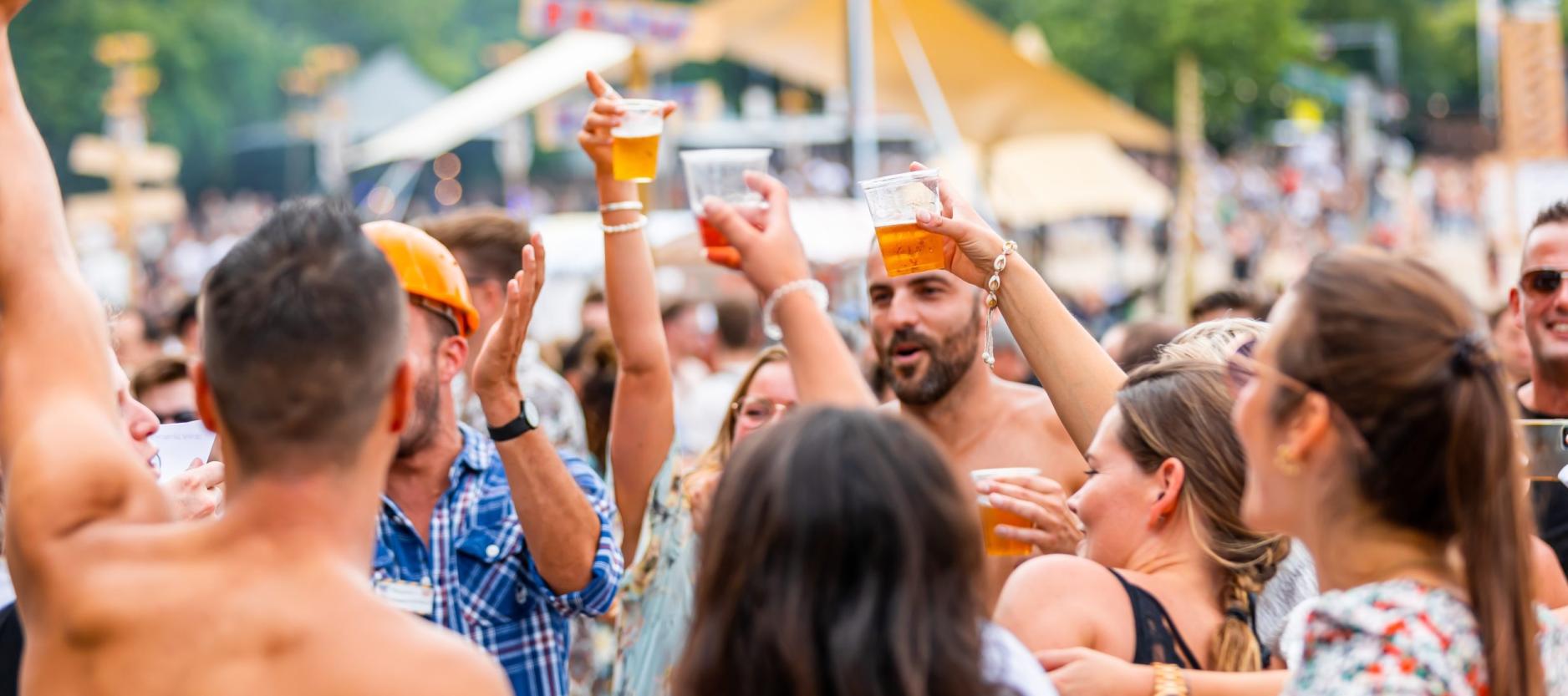 groep jongeren op een festival. handen in de lucht, biertje in de hand. op de achtergrond een grote tent.