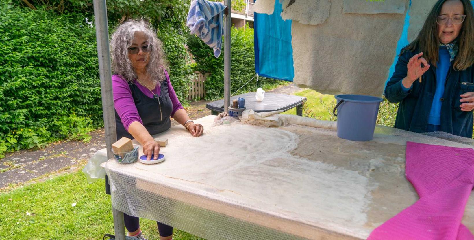 twee vrouwen in een een open tent in de buitenlucht. Groene heg op de achtergrond. Aan een waslijn hangen wollen lappen te drogen. Vrouwen staan een houten tafel. Op de tafel liggen lappen stof en een emmer. Vrouwen zijn bezig met de bewerking van de stof.