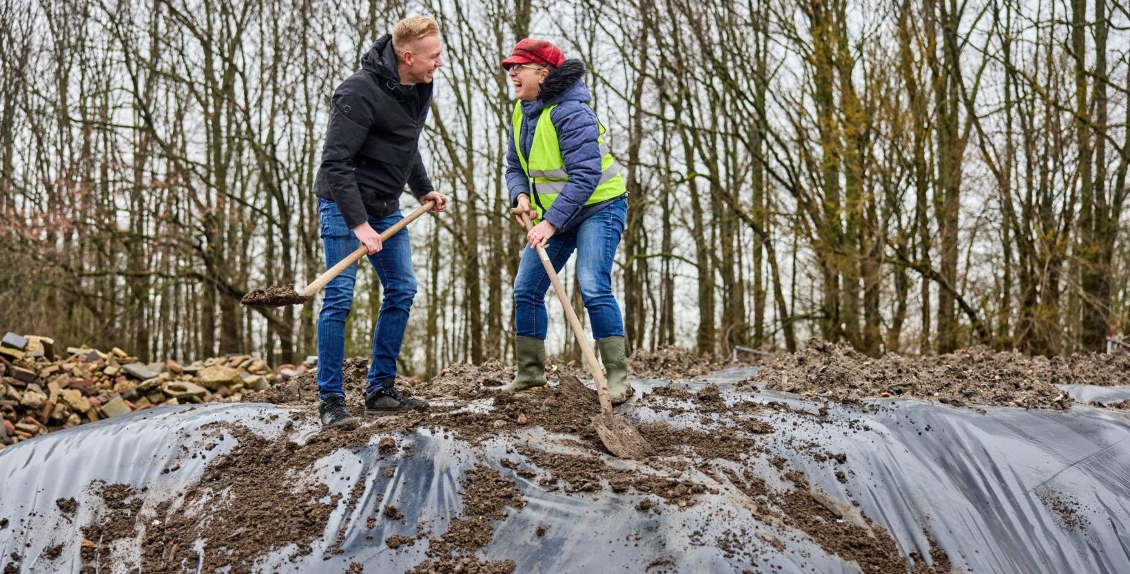 Op de foto twee personen die lachend op de bokashi hoop staan. Beiden hebben een schep in hun handen.