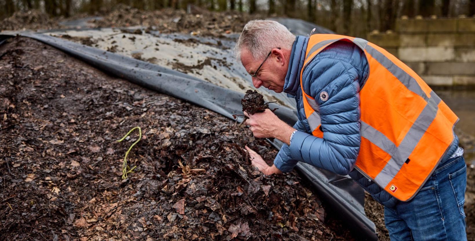 Op de foto een man die met een hand voorover leunt tegen de bokashi hoop. In zijn andere hand houdt hij wat bokashi beet waar hij aan ruikt.